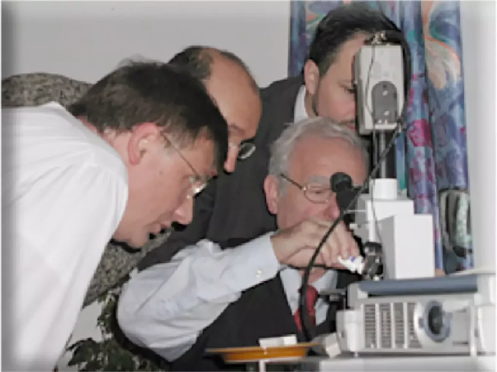 Vier Männer in Hemden und Anzügen blicken konzentriert auf ein medizinisches Gerät in einem Raum mit buntem Vorhang.