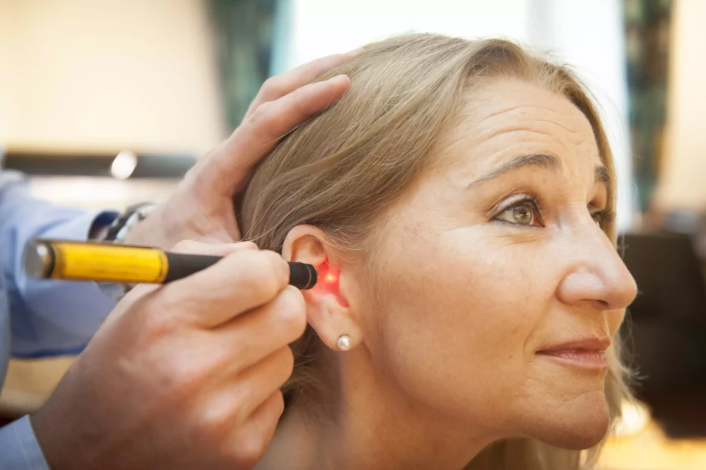 Frau erhält sanfte Ohrbehandlung mit rotem Lichtgerät durch eine Person im hellen Raum, Nahaufnahme seitlich