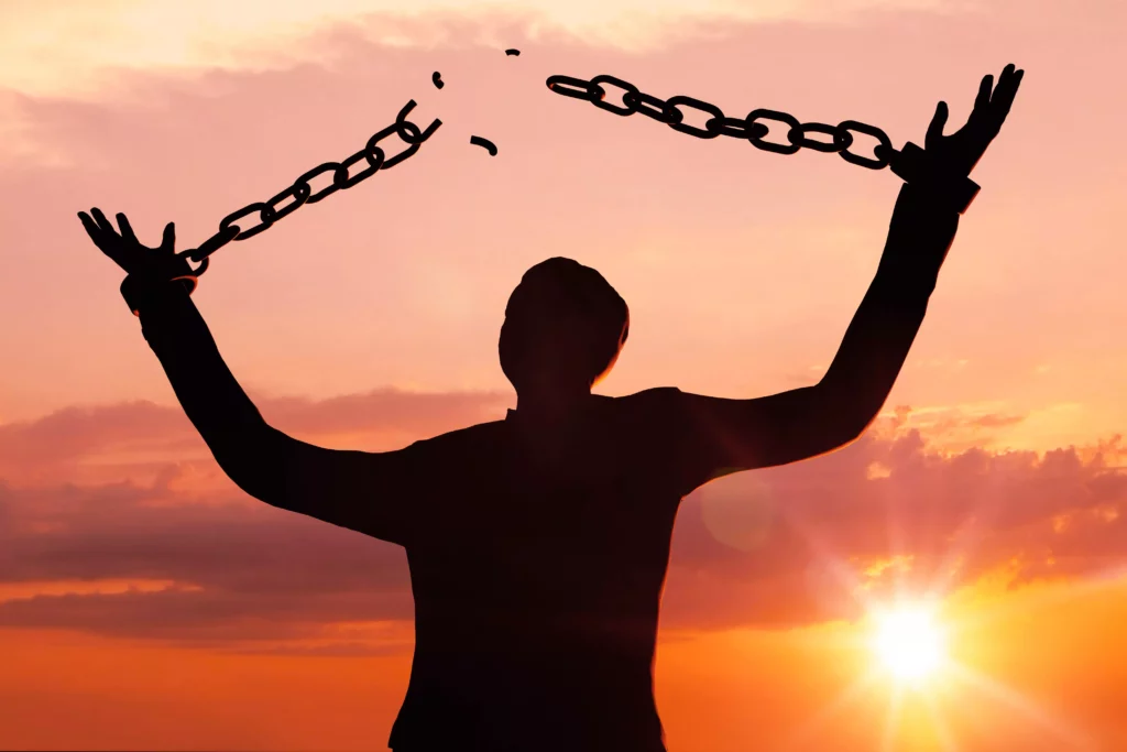 Silhouette einer Person, die sich bei Sonnenuntergang von zerbrochenen Ketten befreit und die Arme erhoben hat