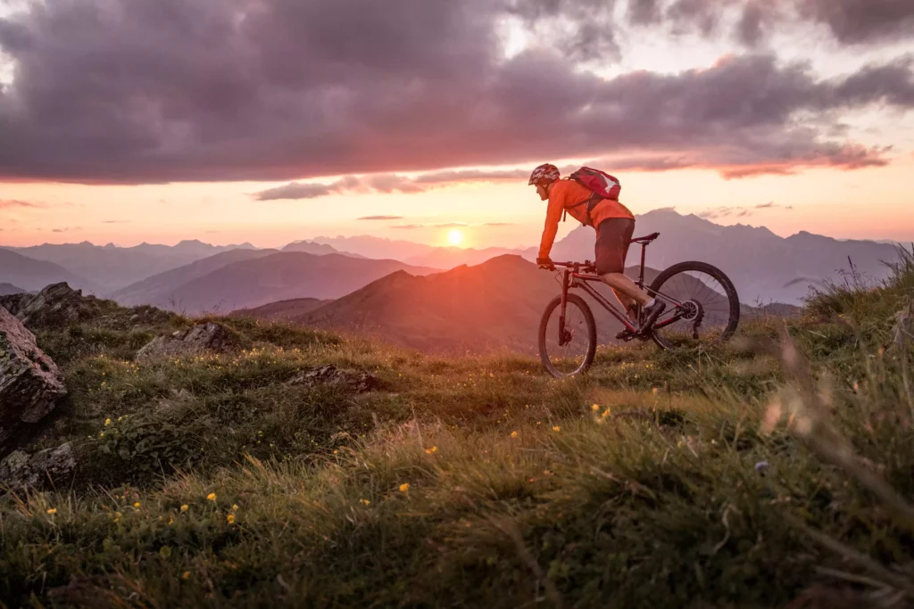 Mountainbiker in orange Jacke fährt bei Sonnenuntergang auf einem Bergpfad mit Blick auf Berglandschaft und Wolkenhimmel