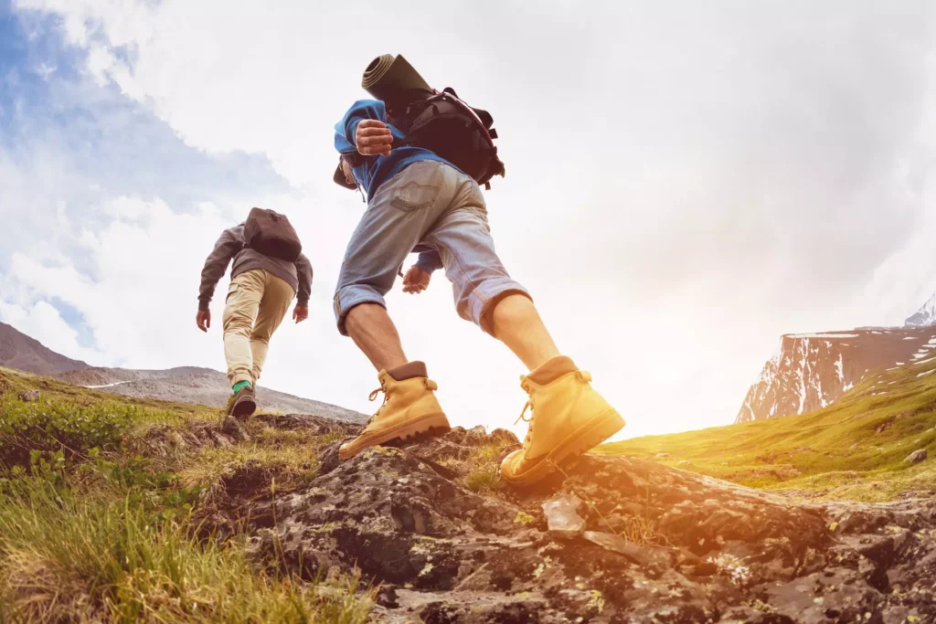 Zwei Wanderer mit Rucksäcken steigen bei Sonnenlicht einen felsigen Bergpfad in einer alpinen Landschaft hinauf.