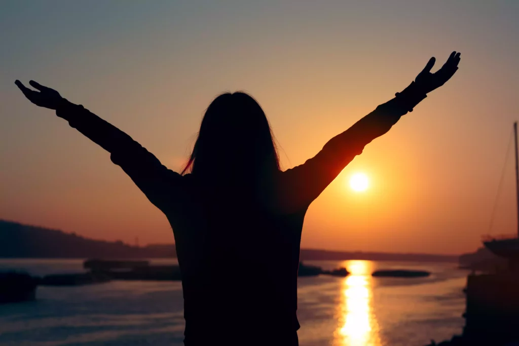 Silhouette einer Frau mit ausgebreiteten Armen am Meer bei Sonnenuntergang, entspannte Urlaubsstimmung am Wasser