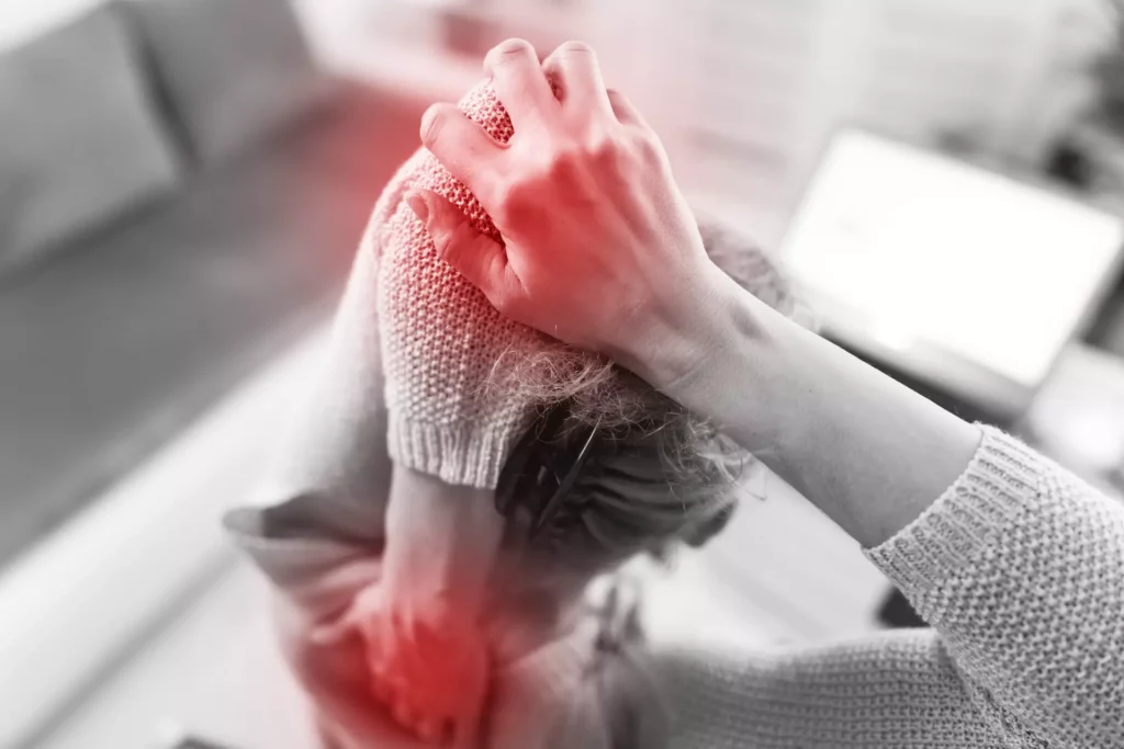 Frau hält sich mit beiden Händen den Nacken und Rücken vor einem Laptop, rote Markierung für Schmerzen sichtbar.