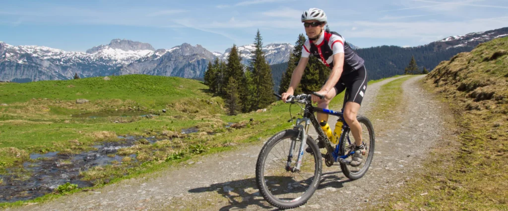 Mann mit Helm und Sonnenbrille fährt Mountainbike auf einem Bergweg mit grünen Wiesen und schneebedeckten Bergen im