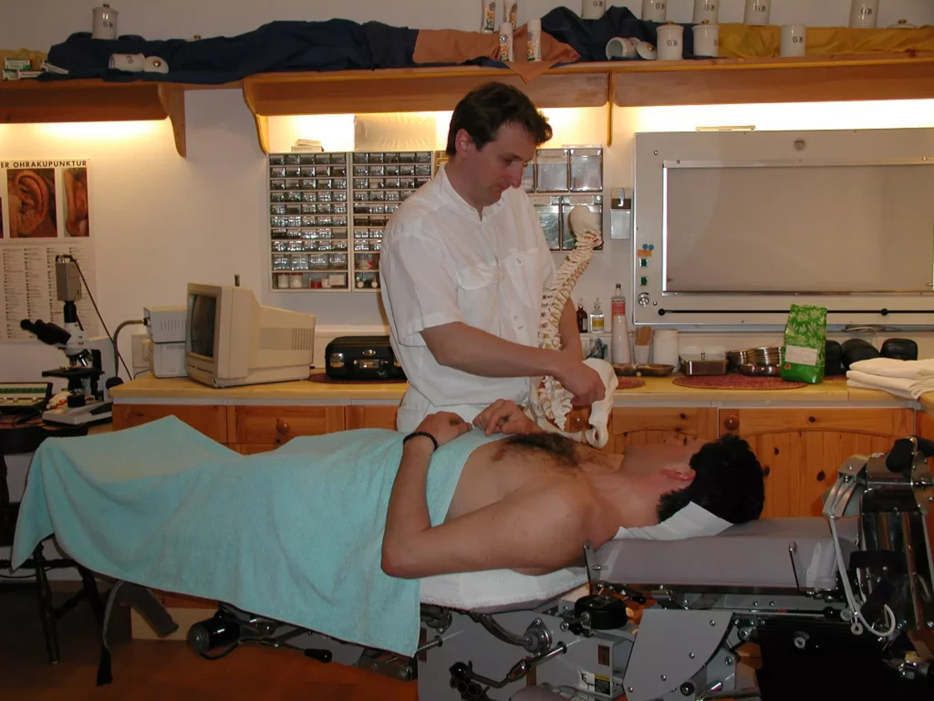 Chiropraktiker in Praxis zeigt Wirbelsäulenmodell einem Patienten auf Behandlungsliege mit Handtuch bedeckt