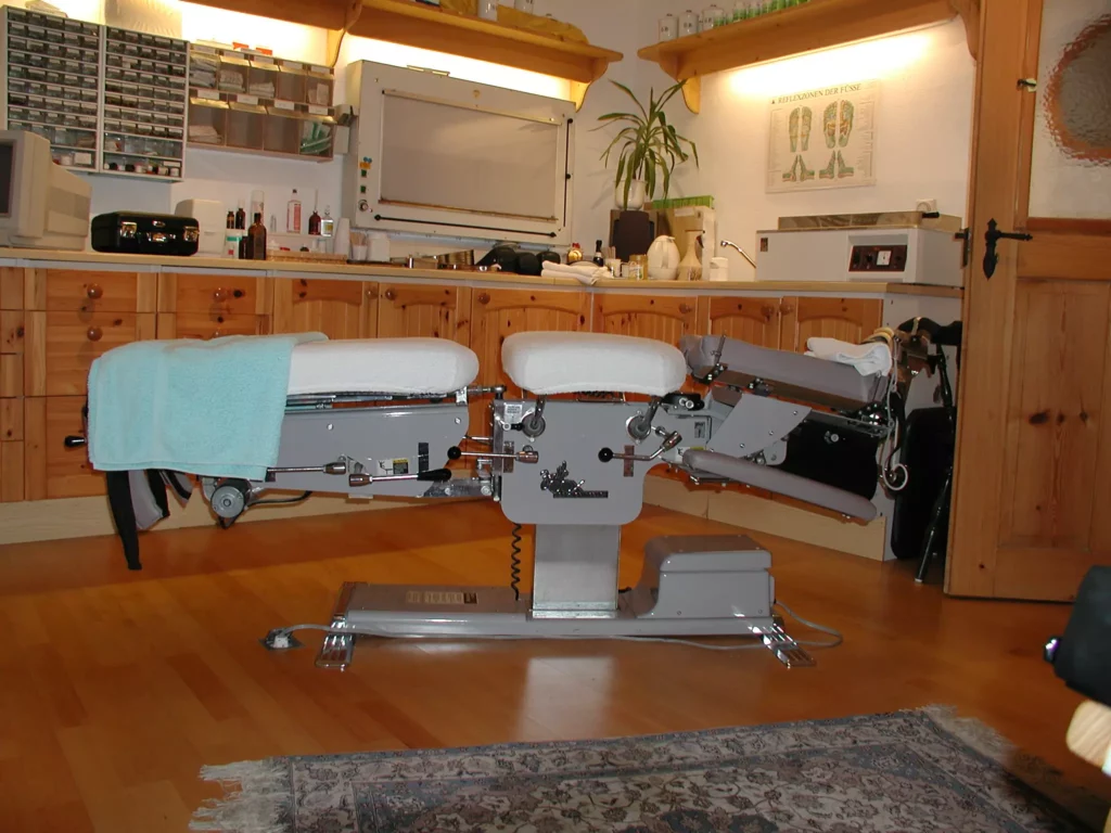 Chiropraktik-Tisch mit Polster und Handtuch in Praxisraum mit Holzschränken und Reflexzonen-Fußposter an der Wand