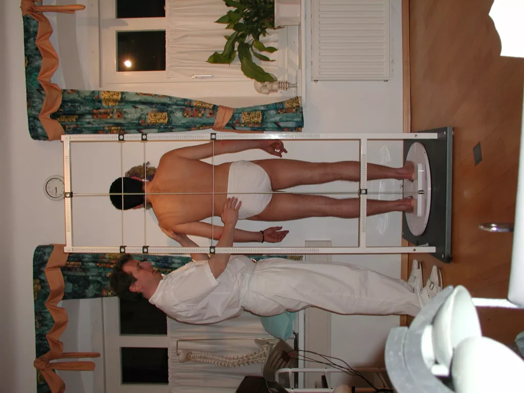 Chiropraktiker misst Wirbelsäule eines Patienten in Unterwäsche mit einem Rastergerät in Praxisraum mit Holzfußboden und