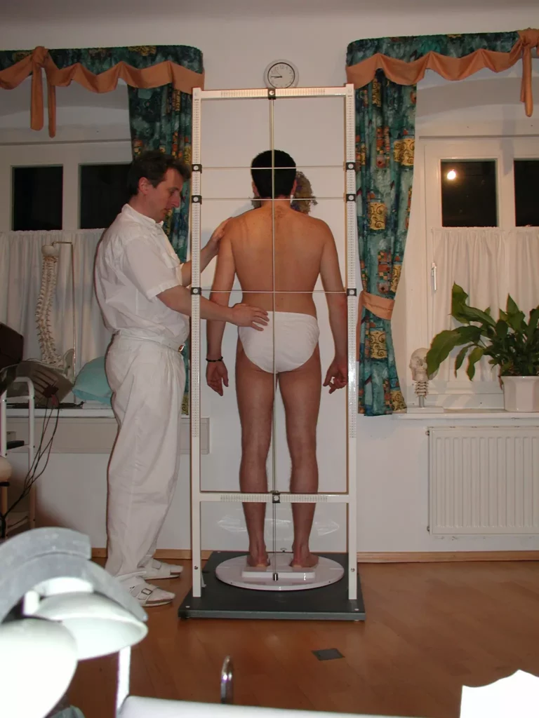 Chiropraktiker misst Wirbelsäulenhaltung eines Mannes mit Spinalyser in Praxis mit Holzfußboden und Vorhängen